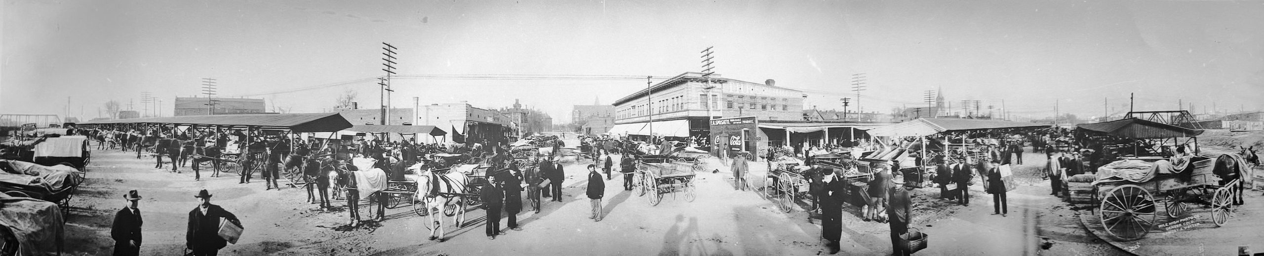 Panoramic black and white photo of Denver’s Denargo Market early in the 1900s, with horse-drawn wagons, produce vendors, and city buildings in the distance.