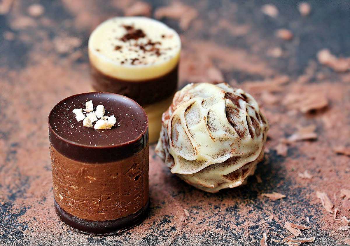 Close-up photograph of three finished chocolates arranged on a dark, cocoa-dusted surface. One cylindrical piece has a glossy dark chocolate top with small pale garnish pieces, another has a light-colored top dusted with cocoa, and a round truffle is covered in a marbled white-and-dark chocolate coating. Image by NoName_13 from Pixabay, used under the Pixabay Content License.