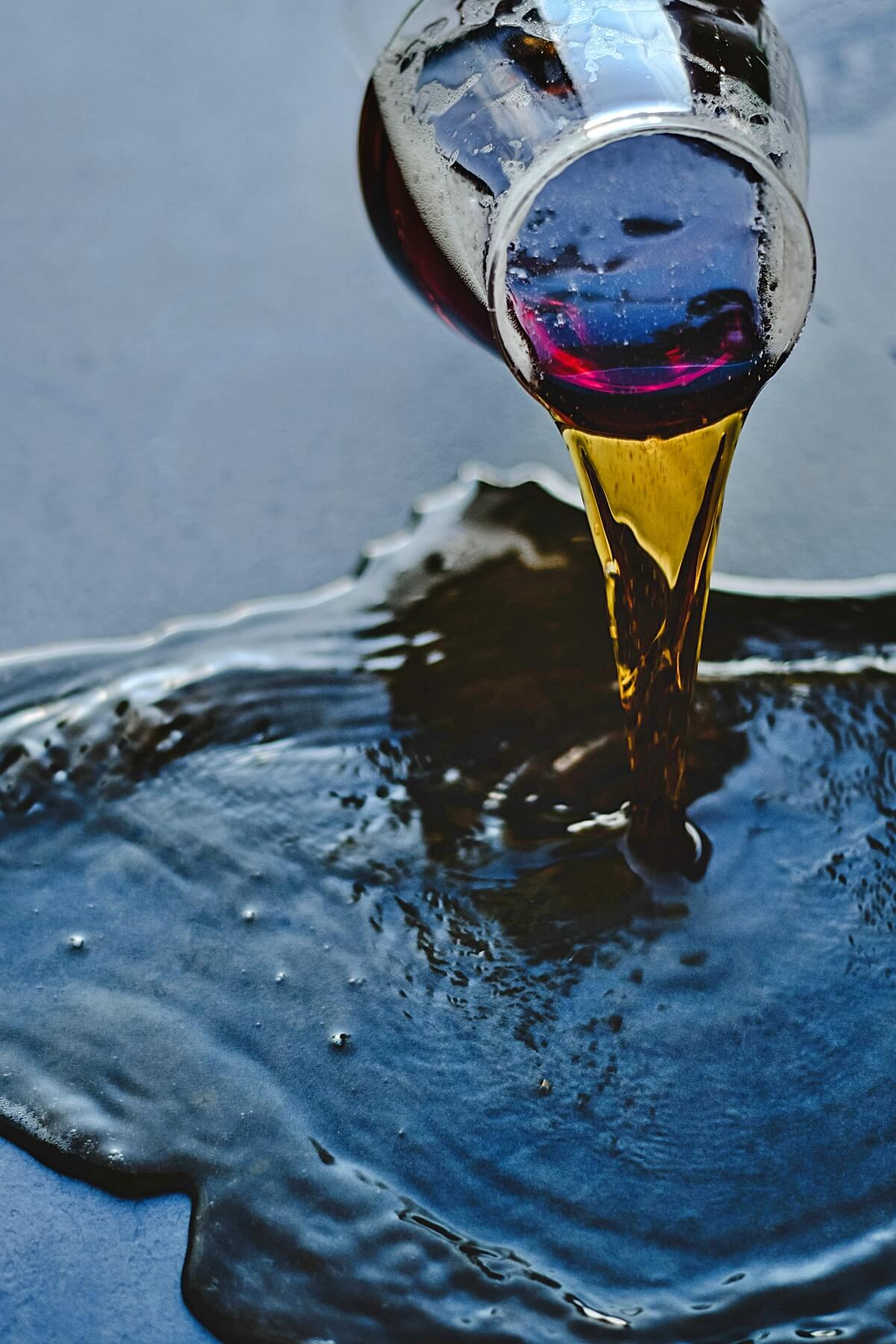 Close-up photograph of dark liquid soy lecithin pouring from a small glass vessel onto a cool blue surface. The stream appears amber-gold where light passes through it, while the spreading pool below reads as deep blue-black with glossy highlights, ripples, and small bubbles. The image emphasizes the ingredient&rsquo;s thickness and fluid movement. Photograph by Eva Bronzini via Pexels.